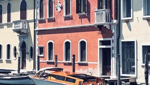 Una hilera de edificios históricos venecianos con ventanas arqueadas y balcones, una embarcación atracada frente a ellos, y cielo despejado.