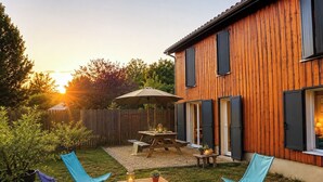 Una casa de madera con un patio, una mesa, sillas y un atardecer de fondo.