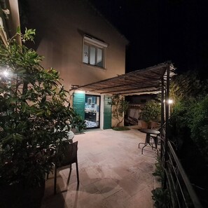 Een overdekt terras met een tafel, stoelen, een bloempot en een toegang tot het huis.
