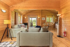 Interior de uma cabana de madeira com sofá, mesa de jantar, luminária e vista pela porta.
