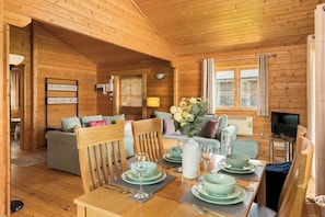 Interior de uma cabana de madeira com uma mesa de jantar posta para quatro, um sofá, uma televisão e uma janela com cortinas.