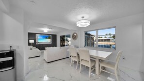 Ein modernes Wohnzimmer mit Flachbildfernseher, weißen Sofas, einem Esstisch mit Stühlen und einem großen Fenster mit Blick auf einen Pool und Palmen.