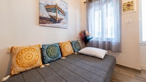 Una habitación con un sofá gris, cojines estampados, un cuadro de un barco, una ventana con cortinas translúcidas y un jarrón con flores secas.