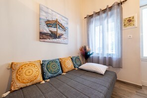 Une pièce avec un canapé gris, des coussins à motifs, un tableau représentant un bateau, une fenêtre avec des rideaux légers et un vase contenant des fleurs séchées.