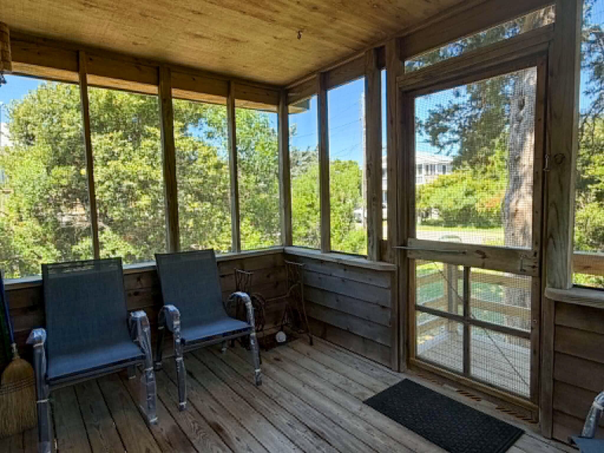 A covered porch with wooden flooring, two metal chairs, and a large window offering a view of trees and a building.