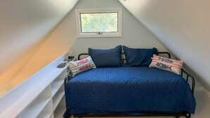 Une petite chambre mansardée de forme triangulaire, avec un lit, des oreillers, une fenêtre et une étagère.