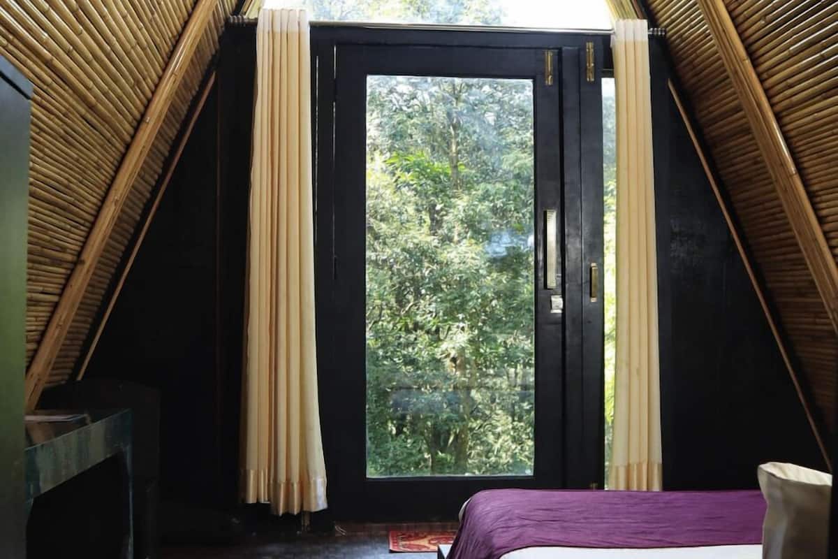 A room with a sloped ceiling, a bed with a purple bedspread, a window with curtains, and a view of trees outside.