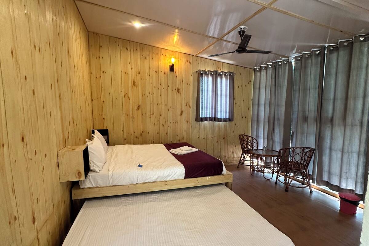 A wooden room with a bed, a table, chairs, a ceiling fan, and a window with curtains.