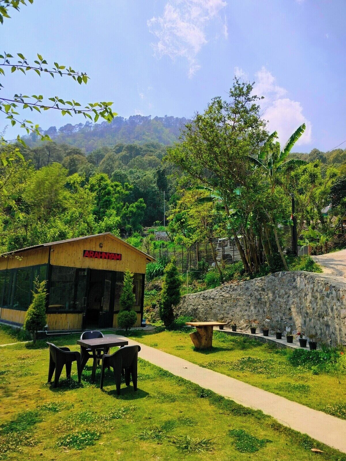 Un bâtiment en bois, sur lequel on peut lire l’inscription « Arahna », un chemin pavé et une végétation luxuriante.. Bhimtal Hotel nr. Bhowali Range Arahnyam
