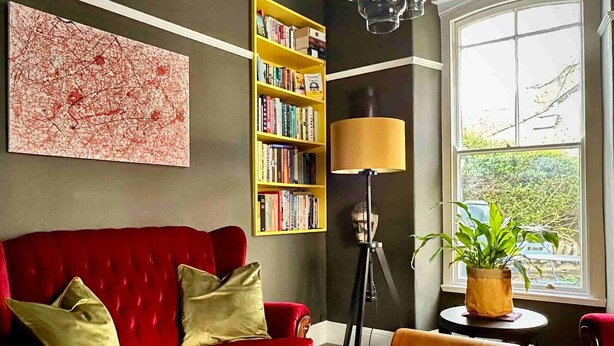 Um sofá vermelho com almofadas douradas, uma estante de livros, um abajur e uma janela com vista para o verde.