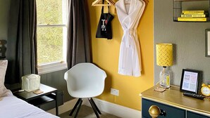 Quarto de hotel com uma cama, uma escrivaninha com abajur, uma cadeira, roupão pendurado em um suporte e uma janela com cortinas.