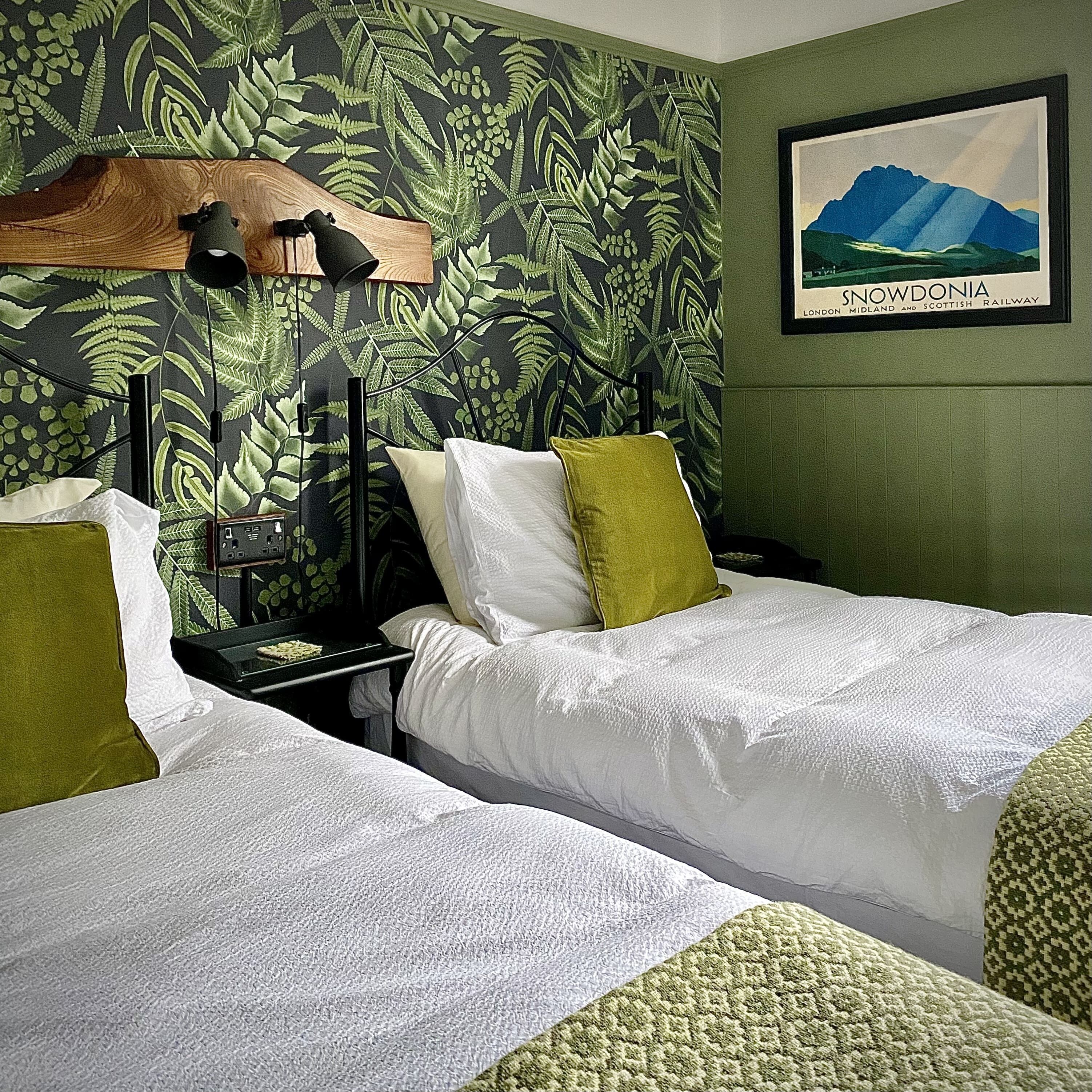 A hotel room with two beds, a nightstand, a lamp, a framed picture of Snowdonia, and a fern-patterned wallpaper.