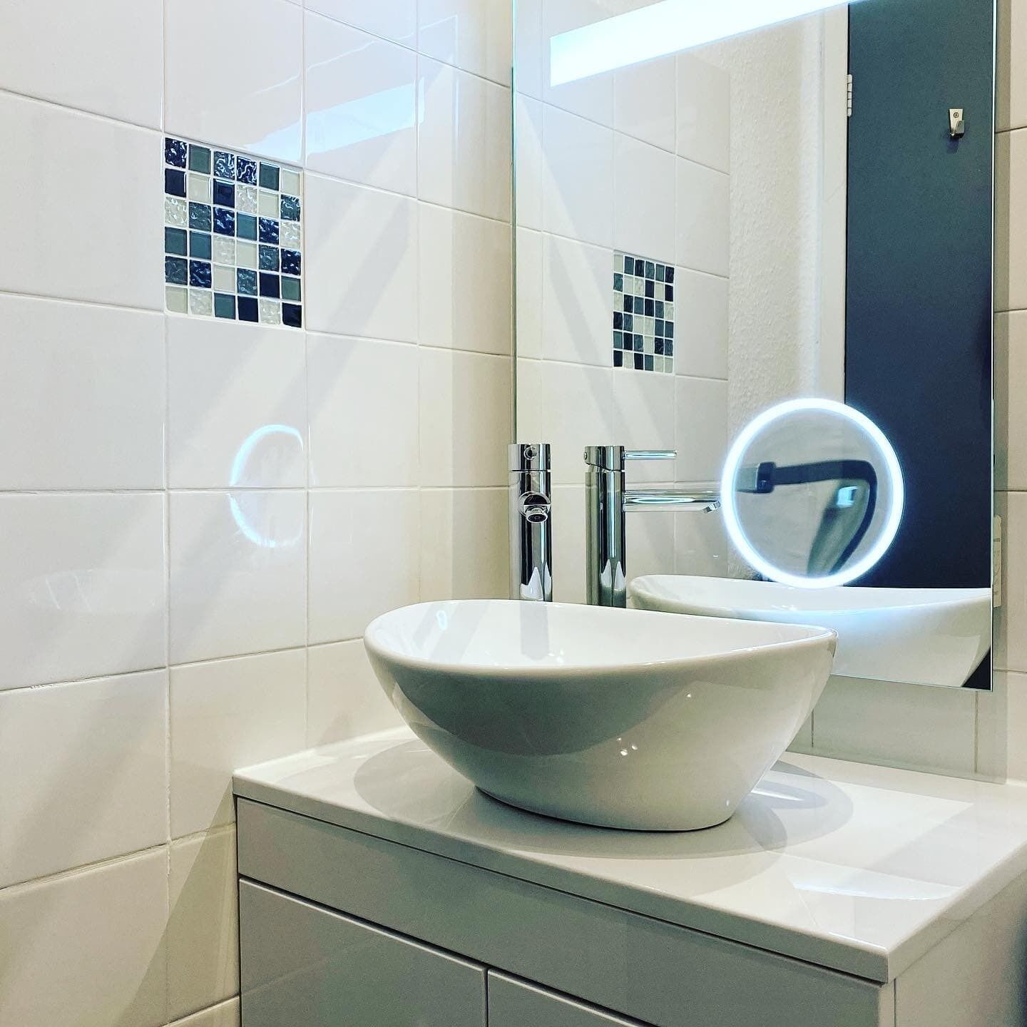 A bathroom with a white sink, chrome faucets, and a mirror with a round light.