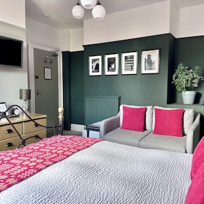 A bedroom with a bed, a sofa, a desk, a lamp, a plant, and framed pictures on the wall.