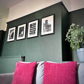Een kamer met een groene wand, een radiator, een grijze bank met rode kussens, en drie ingelijste foto's.