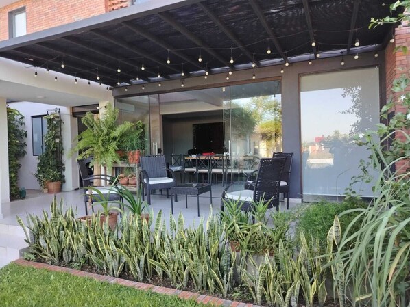 Une terrasse extérieure avec des chaises et des tables, entourée de plantes en pot et d’un mur de verre.