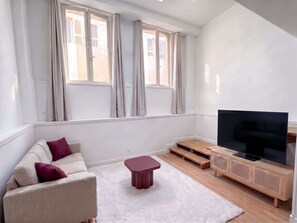 Un salón con sofá, un reposapiés rojo, un televisor sobre un mueble de madera y ventanas amplias con cortinas.