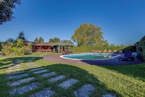 A backyard with a swimming pool, a patio with chairs, and a red building.