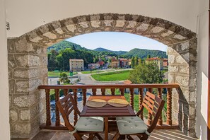 A balcony with a stone arch, a wooden table, two cushioned chairs, and a view of a town with buildings and greenery.