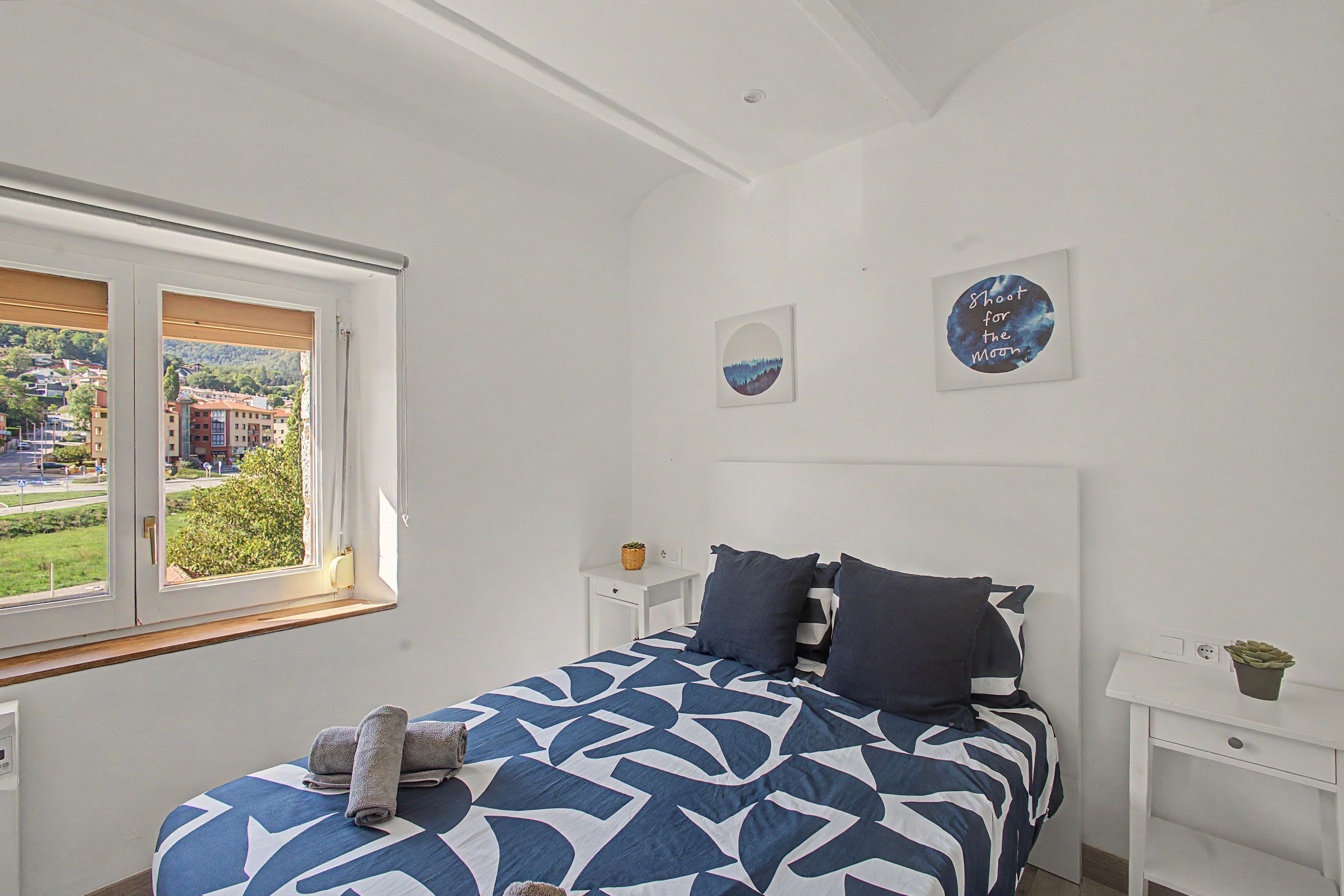 Un dormitorio con cama, mesita de noche, una obra de arte circular en la pared y ventana con vistas a edificios y zonas verdes.