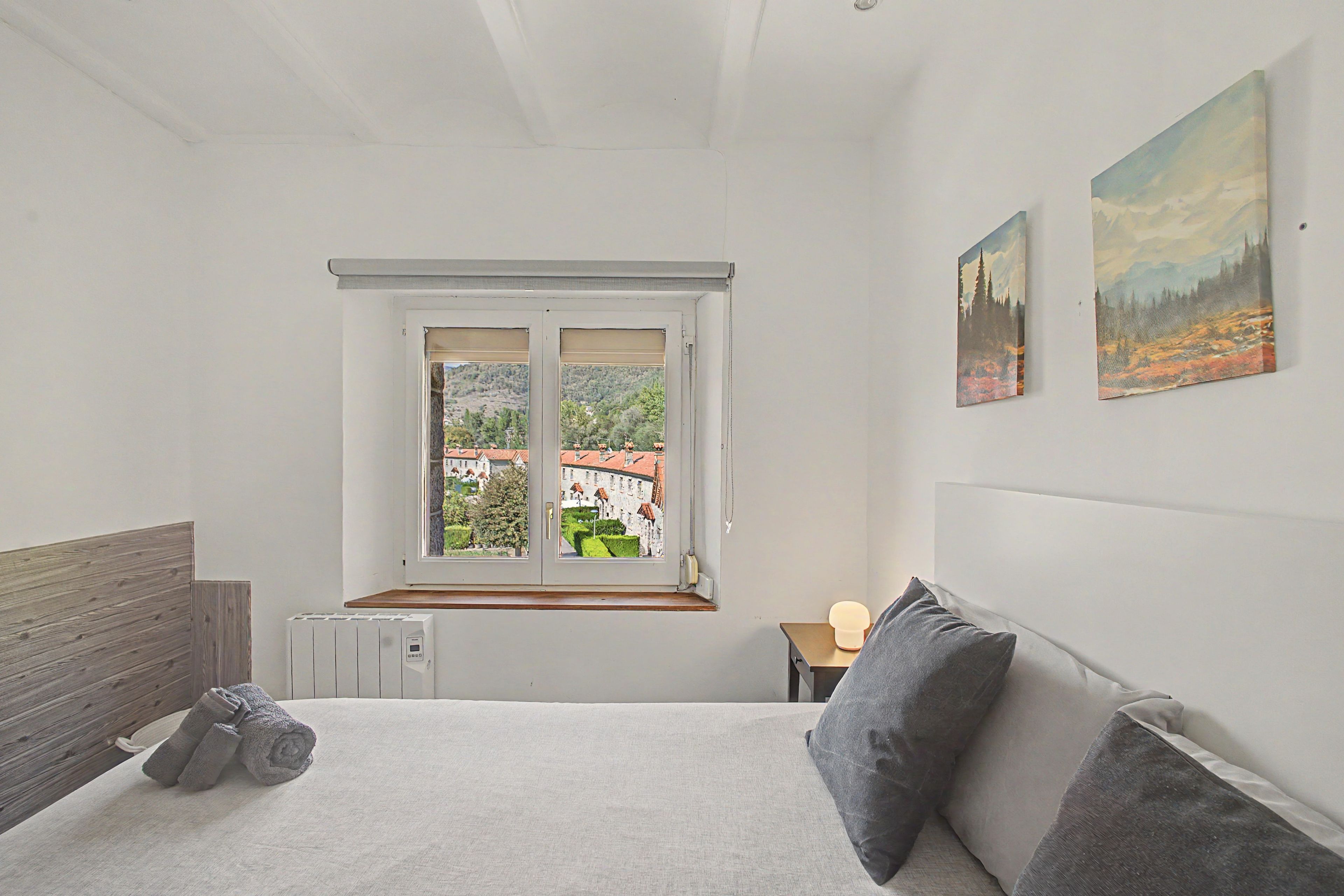 Un dormitorio con cama, ventana con vistas a edificios, radiador y mesita de noche con lámpara.