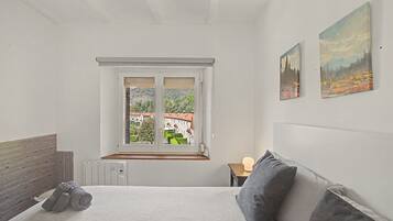 Un dormitorio con cama, ventana con vistas a edificios, radiador y mesita de noche con lámpara.