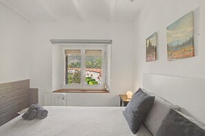 A bedroom with a bed, a window with a view of buildings, a radiator, and a bedside table with a lamp.