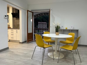 Een eetgedeelte met een ronde tafel en vier gele stoelen, een potplant en uitzicht op een gebouw door een geopende deur.