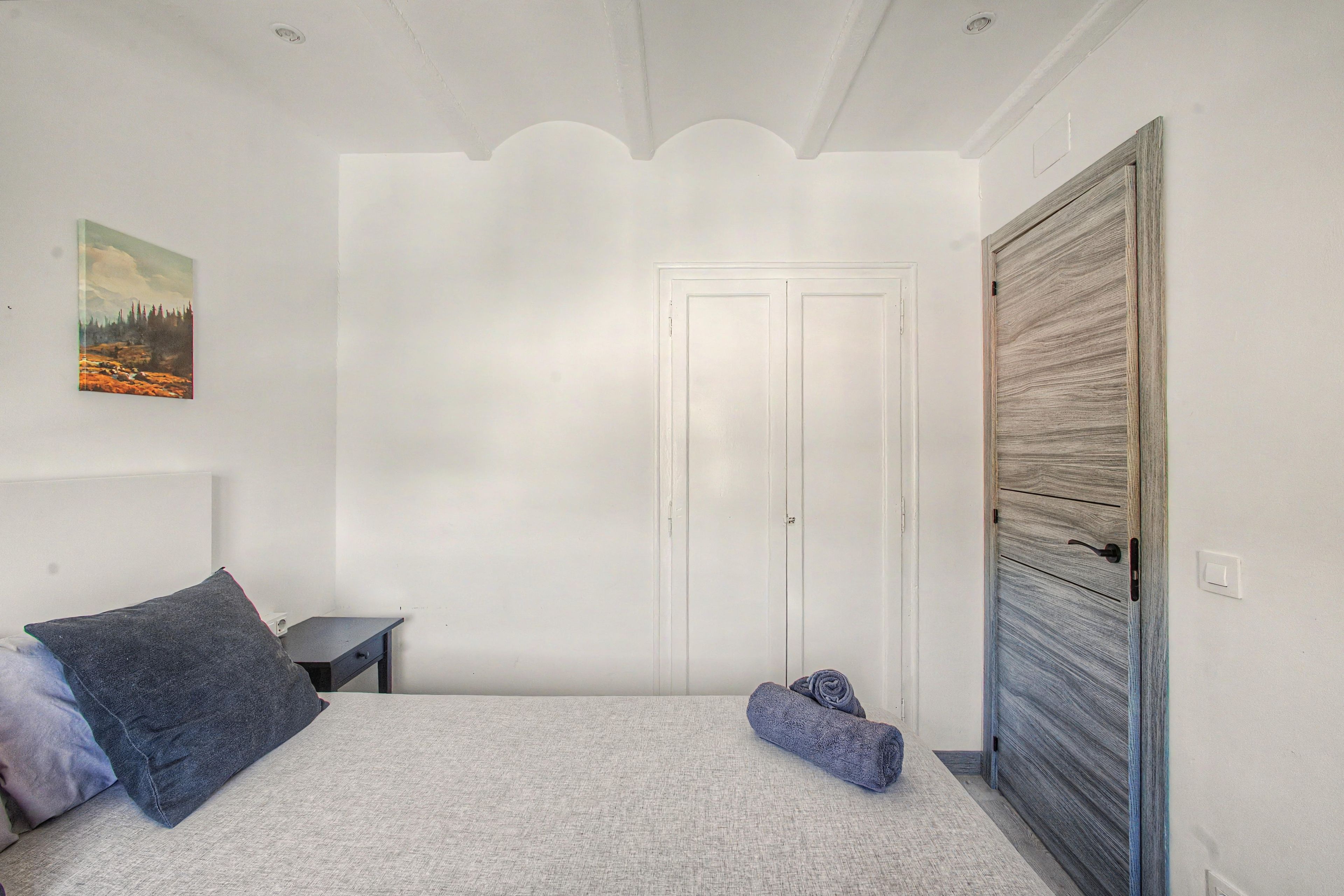 Ein Schlafzimmer mit einem Bett, einem Nachttisch, einer Tür und einem an der Wand hängenden Bild.