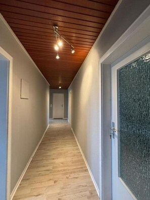 En gang med trægulv, hvide vægge og et loft med træbjælker og indbygget lys.