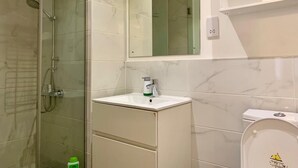 Et badeværelse med en brusekabine med glasvægge, en hvid vask med et spejl over, og et toilet med låg.