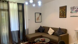 En stue med sofa, sofabord, glastop, lysekrone, gardiner og billeder på væggen.