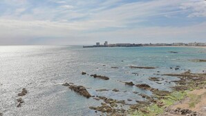 A coastal view with rocky shoreline, calm sea, and distant buildings. - Brasilia - Vue mer d’exception aux Sables, balcon & plage (Les Sables-d´Olonne)