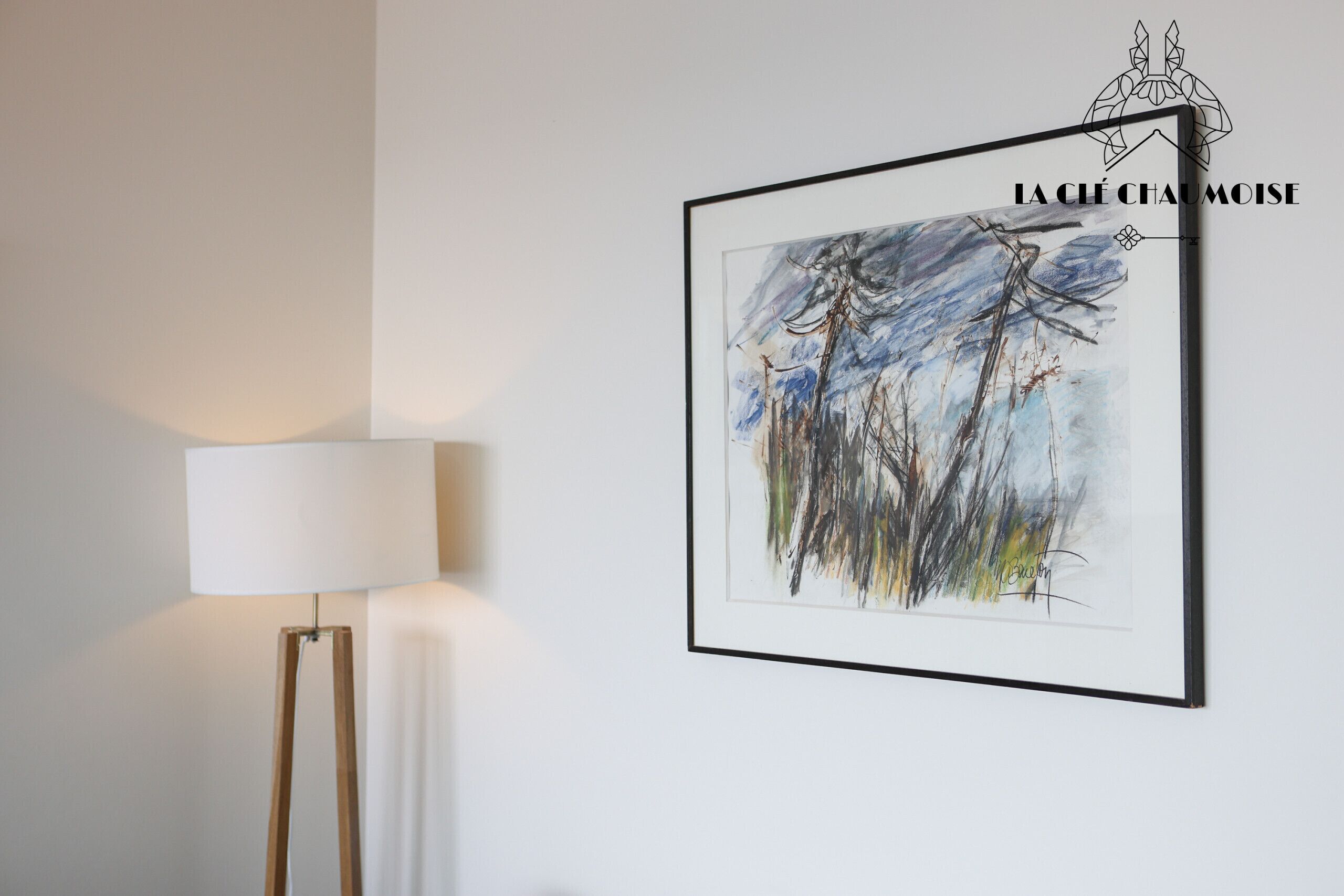 A framed abstract painting on a wall, a floor lamp, and a logo in the top right corner.
