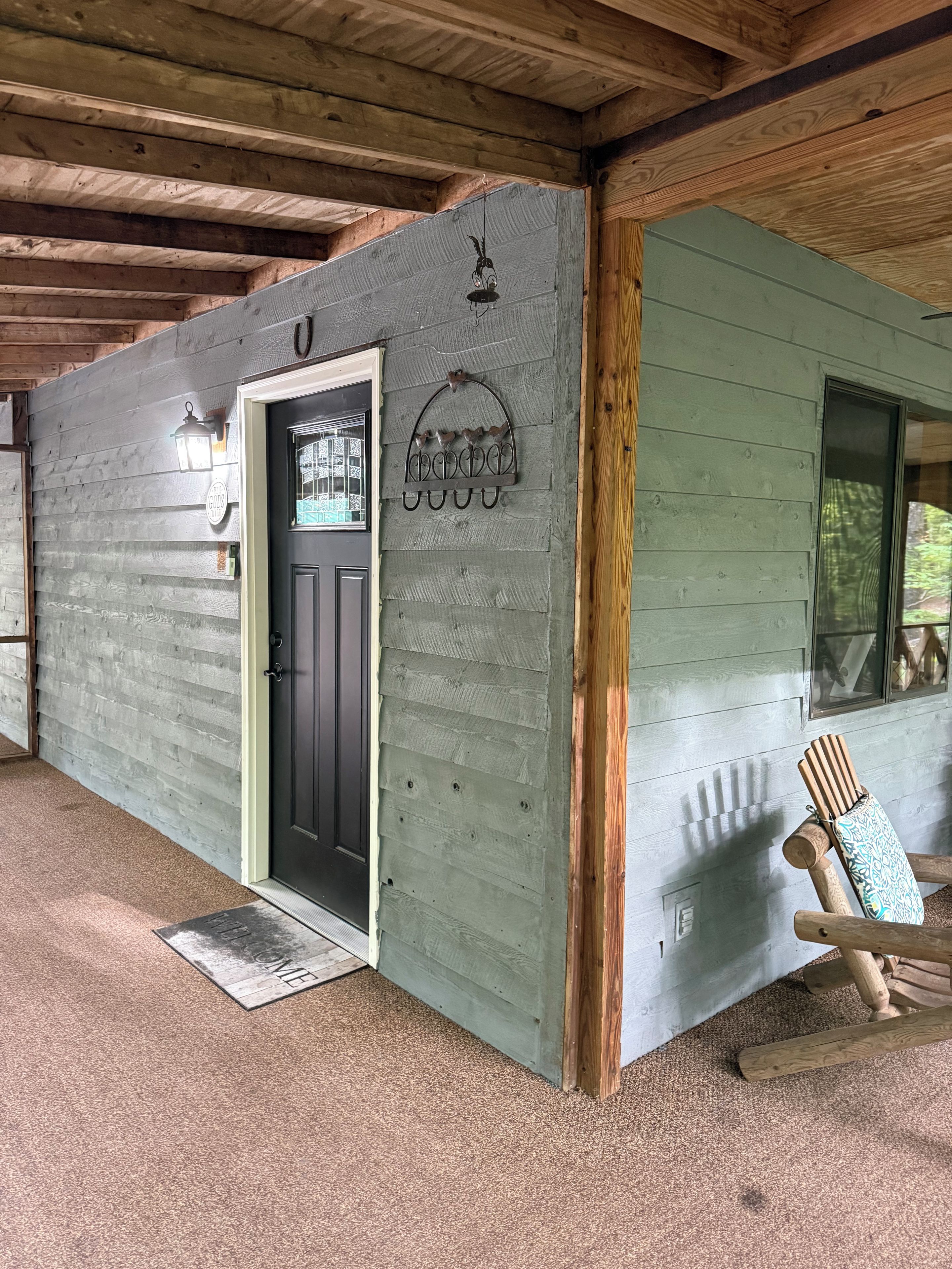 Eine Holzhütte mit einer Holzwand, einer schwarzen Tür, einem Fenster und einer Veranda mit einem Holzstuhl.