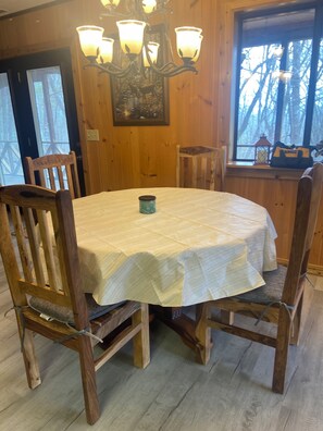 Puisesta ruokapöydästä, jonka päällä on valkoinen pöytäliina, on ympärillä puisia tuoleja. Huoneessa on puuseinät ja kattokruunu.