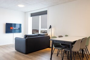 A modern office space with a large flat-screen TV displaying YouTube, a black leather couch, a conference table with chairs, and a window with a city view.