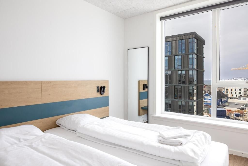 Ein Hotelzimmer mit zwei Betten, einem großen Fenster, einem Spiegel und Blick auf ein modernes Gebäude.