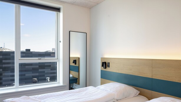 Una habitación de hotel con un ventanal grande, una cama con ropa de cama blanca, un espejo y un cabecero de madera.
