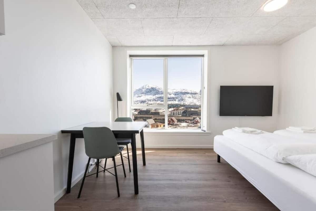 Ett rum med en sÀng, ett skrivbord, en stol, en tv och ett fönster med utsikt över snötÀckta berg.
