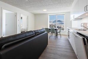 A modern kitchen with a dining area, a black sofa, and a view of a snowy mountain. - Aurora City Apartments (Nuuk)