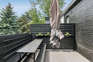 Eine Holzterrasse mit einer Bank und einem hängenden Sonnenschirm, umgeben von einem schwarzen Zaun und viel Grün.