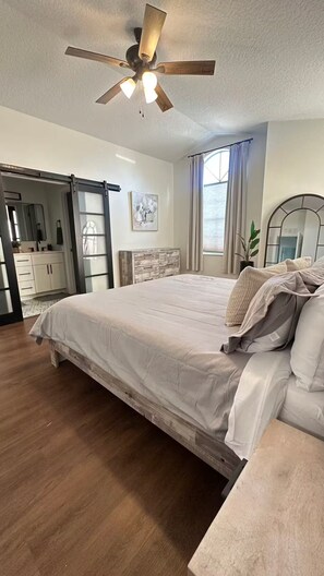 Ein Schlafzimmer mit einem Bett, einem Deckenventilator, einer Kommode, einem Spiegel und einem Fenster mit Vorhängen.