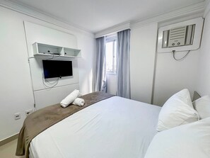 Ett hotellrum med en säng, en tv som är monterad på väggen, ett fönster med gardiner och en väggmonterad luftkonditioneringsanläggning.