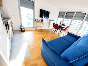 A modern living room with a blue sofa, wooden flooring, a dining area with red chairs, a TV mounted on the wall, and large windows. - Modern & Smart Paris 13 Bibliothèque François Mitterrand (Paris)