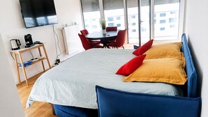 Ein modernes Hotelzimmer mit einem großen Bett, roten und gelben Kissen, einem Fernseher, einer Kaffeemaschine und einem Essbereich mit roten Stühlen.