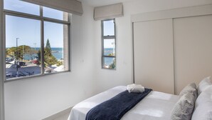 Quarto com uma janela grande, cama com roupa de cama branca e vista para o mar e edifícios.