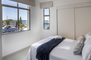 A bedroom with a large window, a bed with white bedding, and a view of the sea and buildings. - Sea Front - 3Q Wonderful and cozy (Vila Velha)