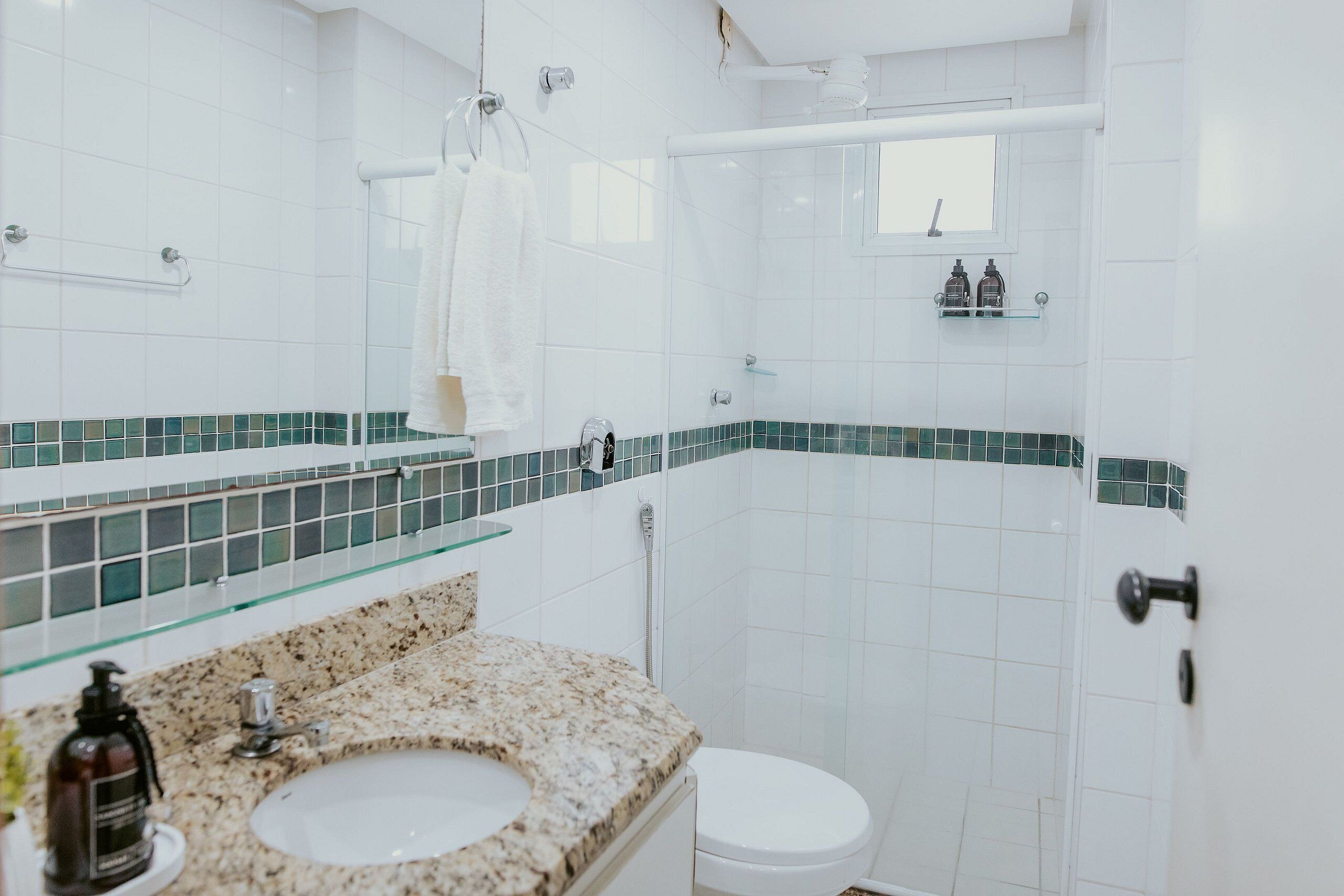 Baño con lavamanos, inodoro, ducha y repisa de vidrio con artículos de aseo.