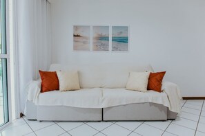 Ein weißes Sofa mit orangefarbenen Kissen, eine weiße Wand und drei Bilder mit Strandmotiven.